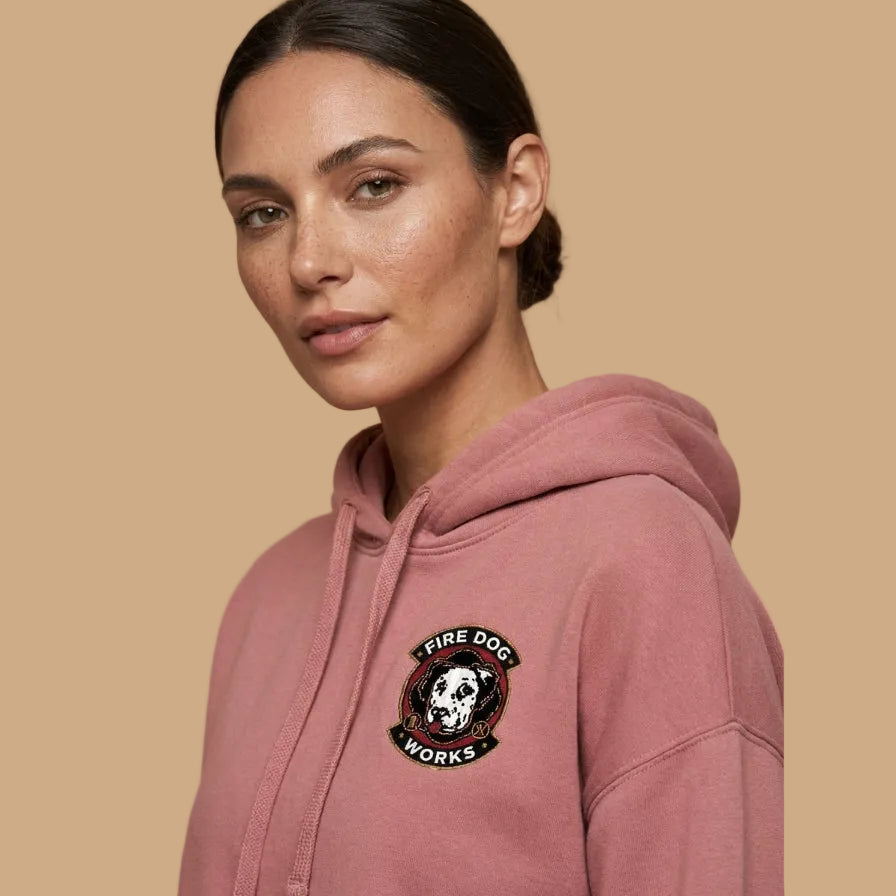 A woman wearing a dusty rose pink hoodie with a circular embroidered patch on the left chest that reads 'FIRE DOG WORKS' around a dog skull graphic, set against a solid beige background.
