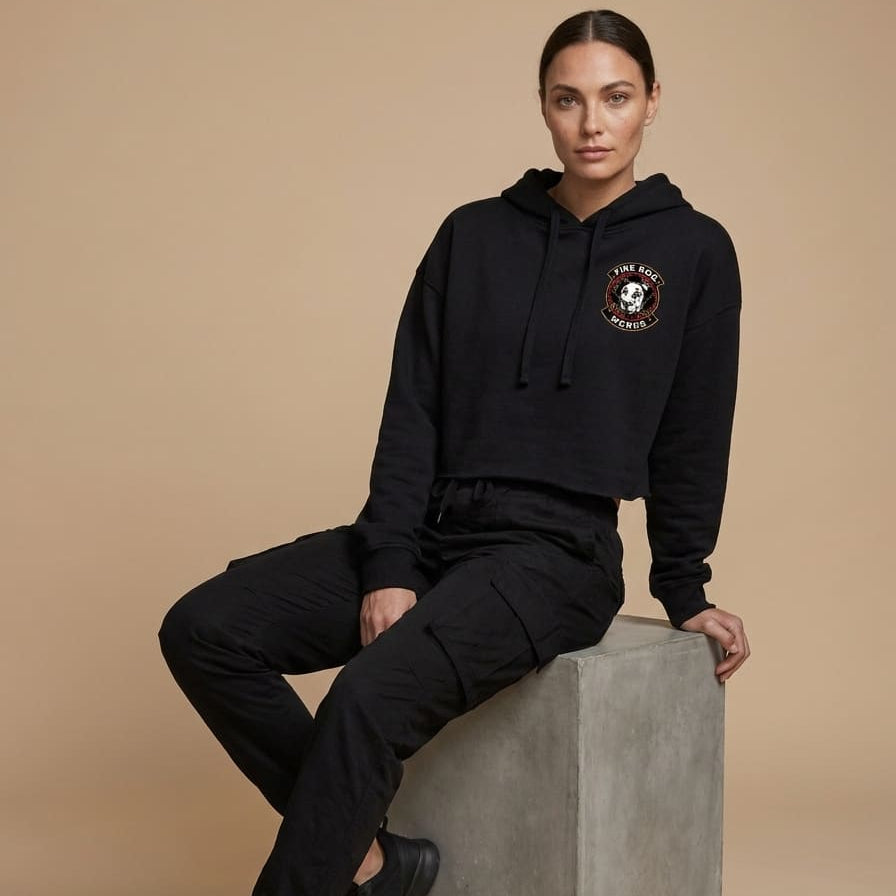 A model wearing a black cropped hoodie with a circular embroidered crest on the left chest, paired with black cargo pants and black sneakers, sitting on a gray concrete block against a solid beige studio background.