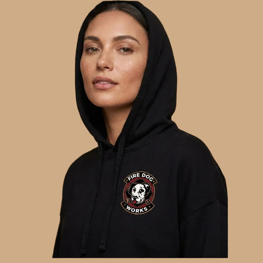 A woman wearing a black pullover hoodie with a circular 'Fire Dog Works' graphic on the left chest, set against a solid beige background.