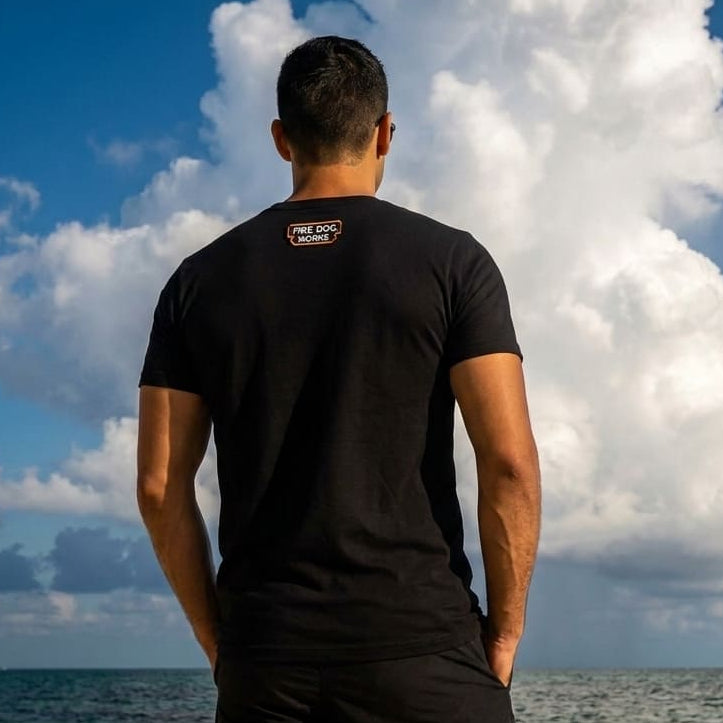 A man stands on a beach with his back to the camera, wearing a black t-shirt and black shorts, looking up at a large white cloud against a bright blue sky. A small 'The Sports Brand' logo is visible on the back of his shirt.